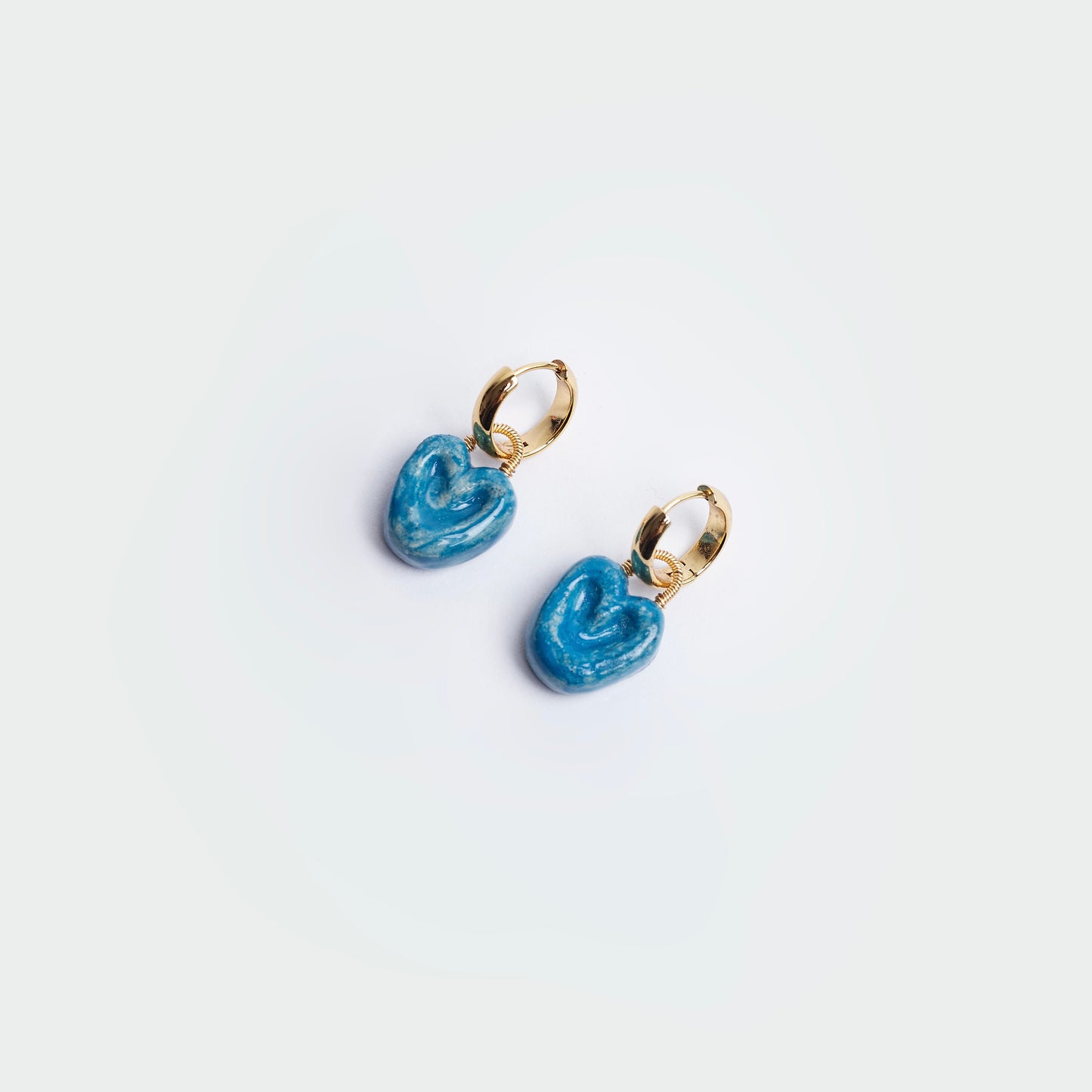 aretes colgantes estilo hoops en fondo blanco, hechos a mano en material de cerámica color azul con candongas en acero inoxidable color dorado.