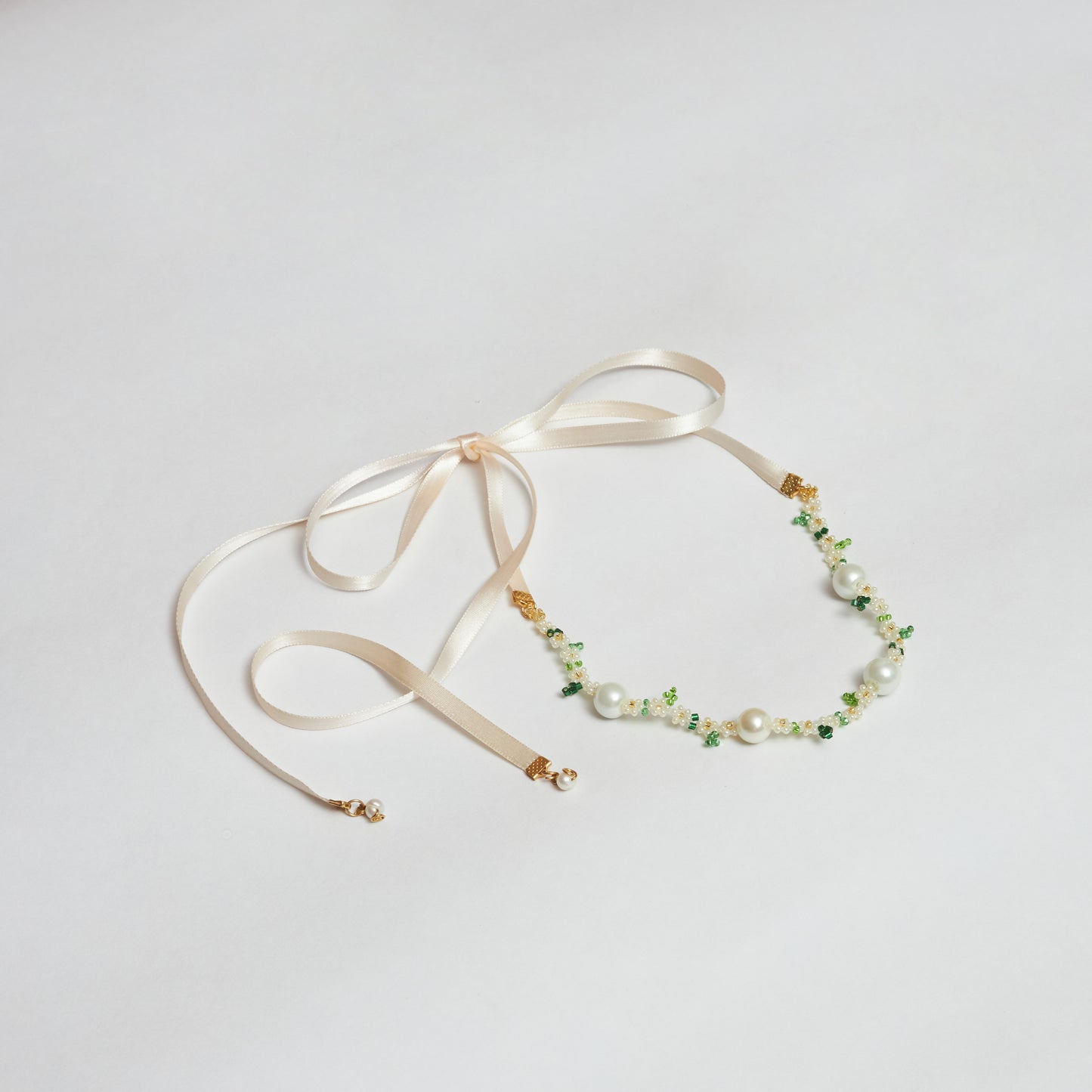 Collar estilo chocker tejido a mano en mostacilla y rocaya con forma de flores en colores verde claro, verde oscuro, blanco y dorado, acompañado con muranos en color blanco, tapanudos y cintillas color rosa pastel atadas en forma de moño.
