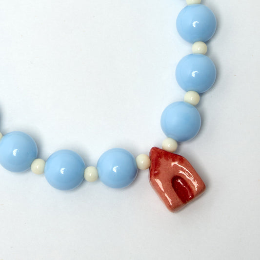 Collar de fiesta estilo chunky hecho con cuentas color celeste y perlas color marfil  con un dije en forma de casa en material de cerámica porcelana con colores rojo y ladrillo.
