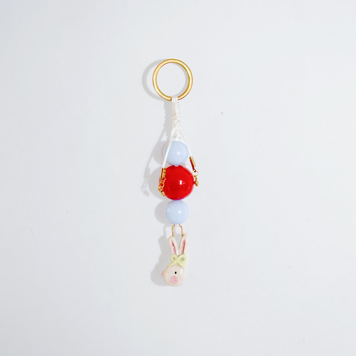 Llavero estilo charm bag con cuentas en color celeste y rojo y un dije colgante en cerámica con forma de conejo coquette en colores blanco, rosado y verde atados en vertical.