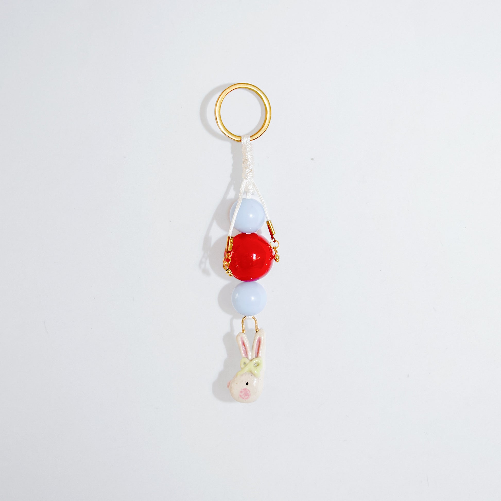 Llavero estilo charm bag con cuentas en color celeste y rojo y un dije colgante en cerámica con forma de conejo coquette en colores blanco, rosado y verde atados en vertical.