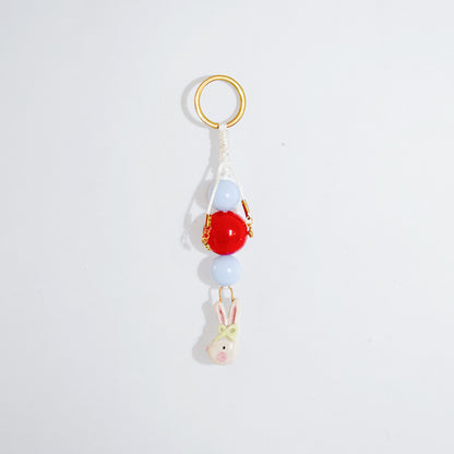 Llavero estilo charm bag con cuentas en color celeste y rojo y un dije colgante en cerámica con forma de conejo coquette en colores blanco, rosado y verde atados en vertical.