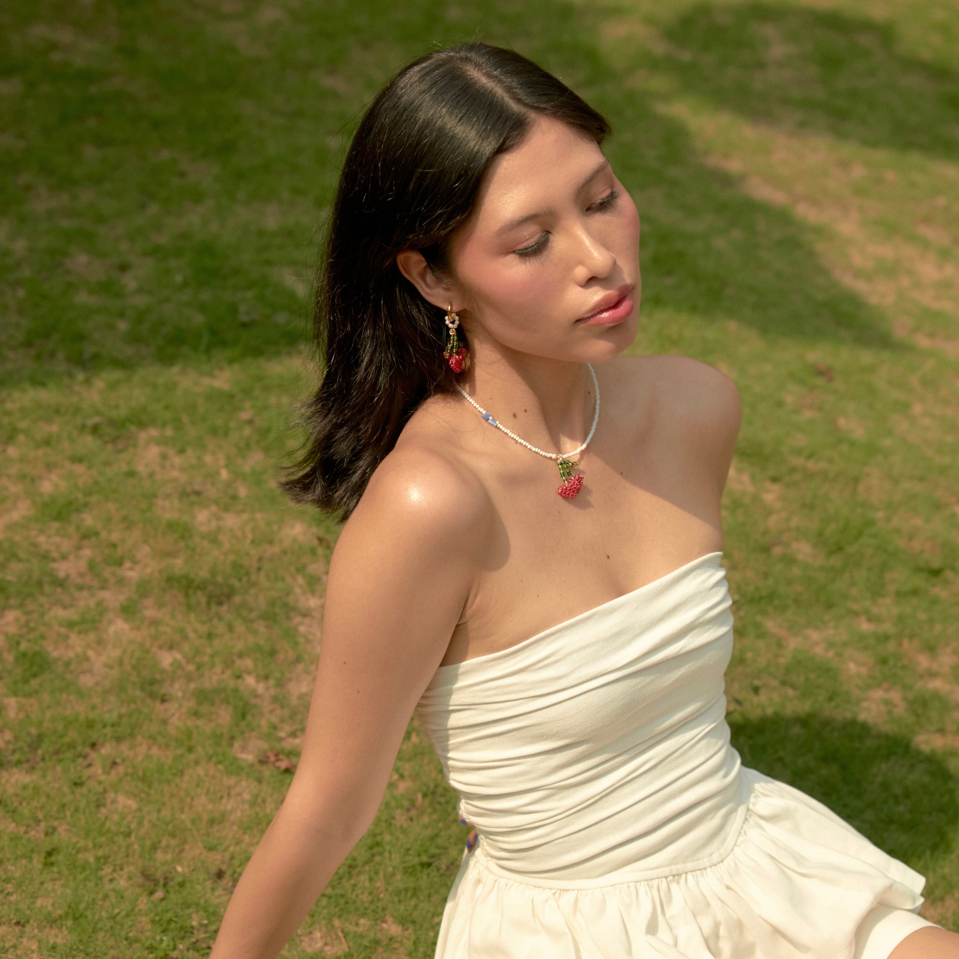 Modelo de tes blanca y cabello negro, usando un vestido blanco y juego de accesorios de collar y candongas con dijes en forma de cerezas de corazón tejidas a mano